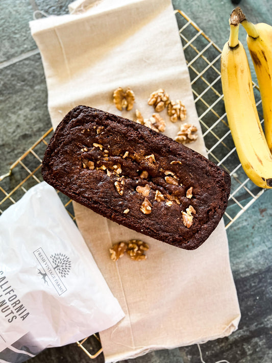 A loaf of freshly baked gluten free banana bread on a cooling rack with a bunch of 2 bananas to the right and a bag of Fresh Vintage Farms raw california grown walnuts to the left